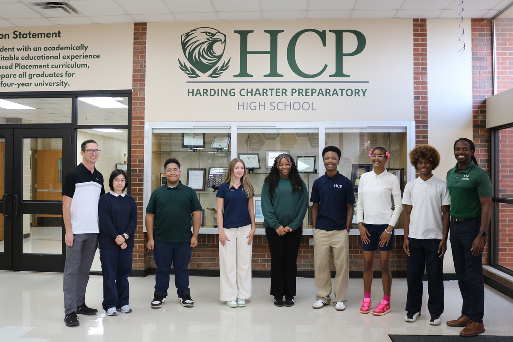 Nine people, including adults and students, stand outside a school entrance. The sign reads "Harding Charter Preparatory High School."