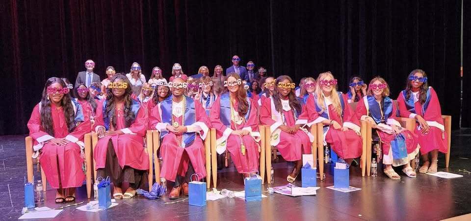Edgewood High School's 2026 International Baccalaureate graduates seated on stage in red and blue graduation gowns, wearing celebratory "2026" novelty glasses, with gift bags at their feet.