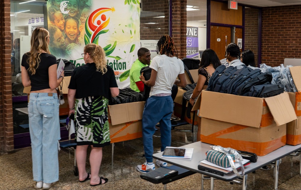 A group of people gather around tables lined with cardboard boxes full of backpacks, sorting and distributing them in a school hallway. An "Elee Foundation" banner with smiling children hangs on the wall behind them