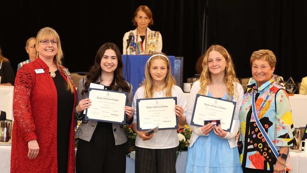 Five women stand in front of another woman at a podium. Three of them are young ladies or students and are holding certificates
