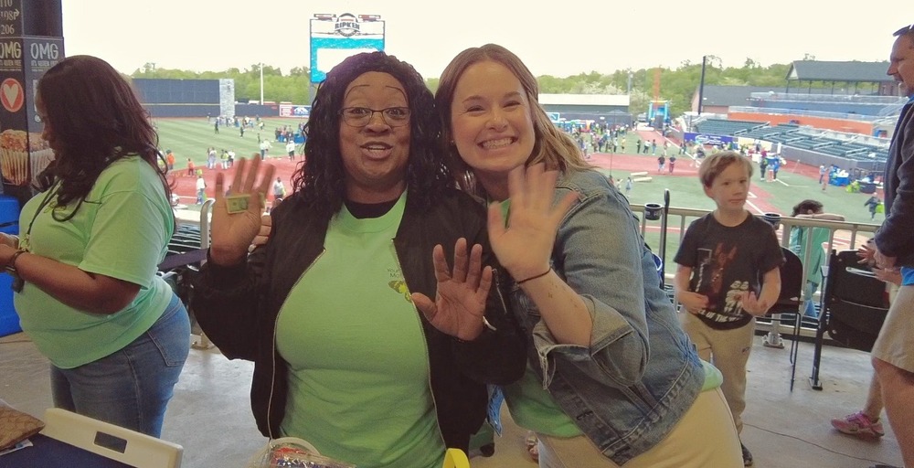 Two HCPS employees standing at a table in front of a baseball field smiling and waiving
