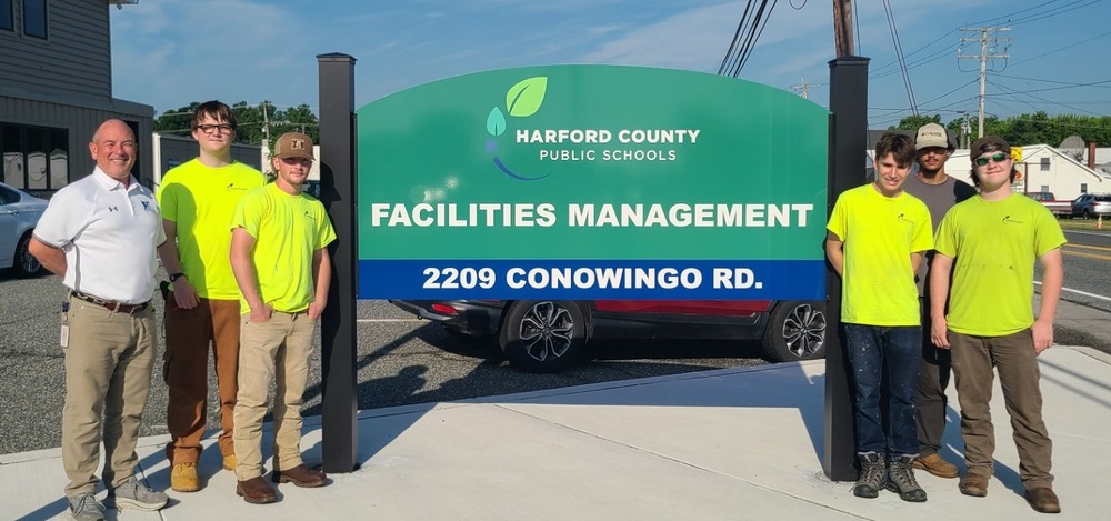 Five students and an adult stand in front of the Facilities Managerment sign
