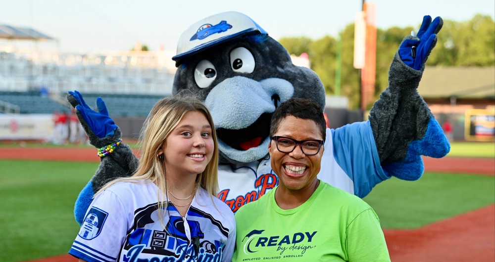 Dr. Mack wearing a green shirt standing with a young woman in an iron birds uniform with the ironbirds mascot standing behind them.