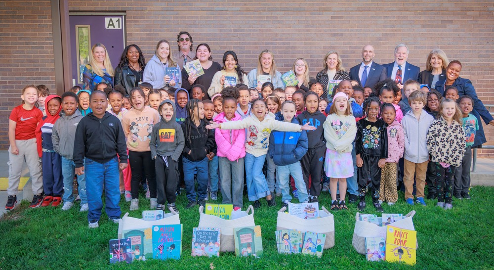 A group of students and adults standing outside a building smiling and showing books that were donated