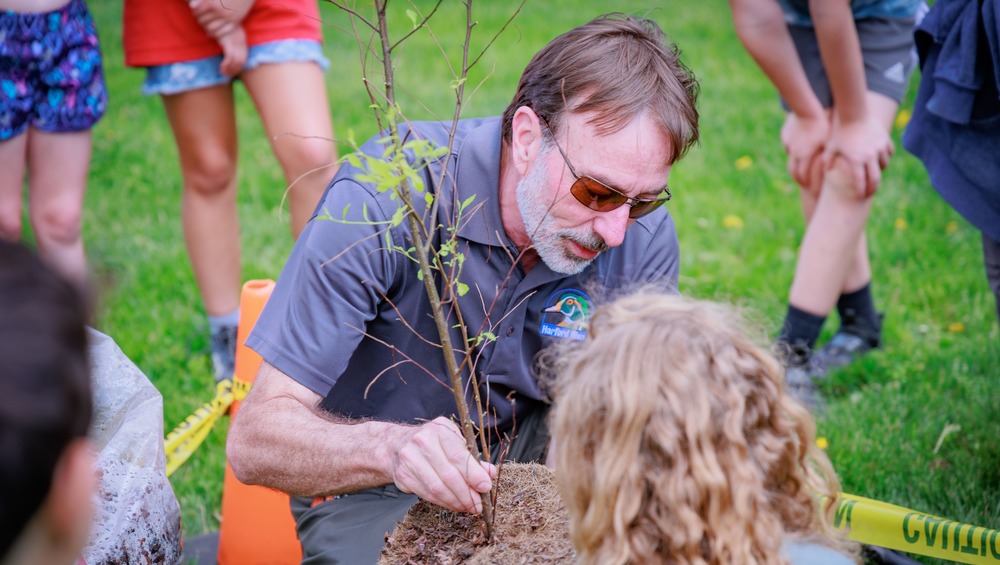 Howard Eakes helps a student hold a sapling while they plant it in the ground