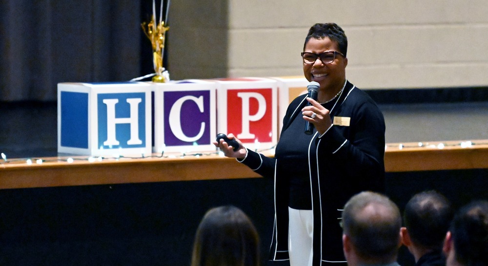 Dr. Mack standing in front of a group of educators and a sign that says HCPS smiling into a microphone