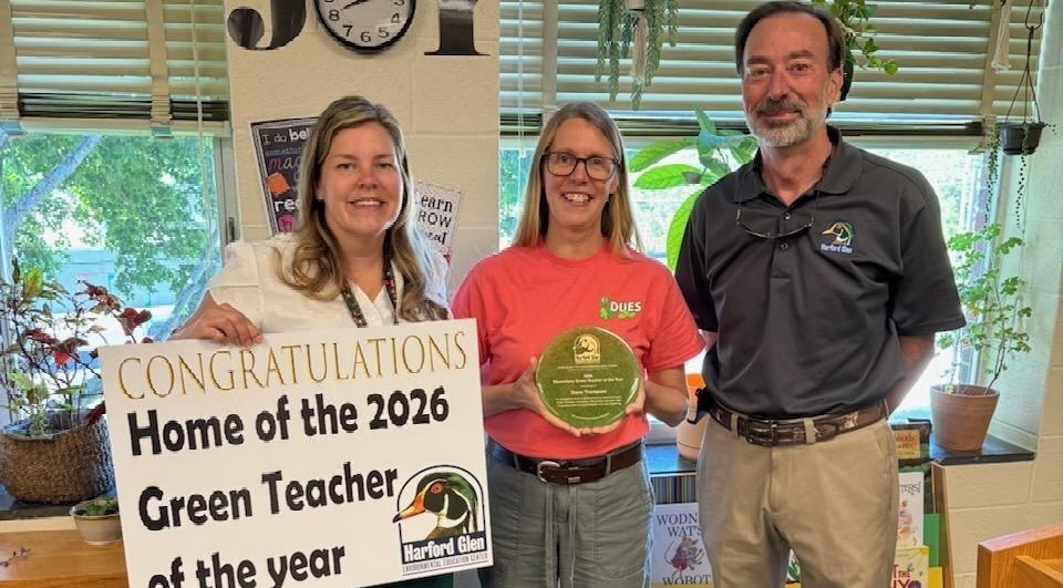 Three people in a classroom with one holding a sign that says congratulations home of the 2026 green teacher of the year