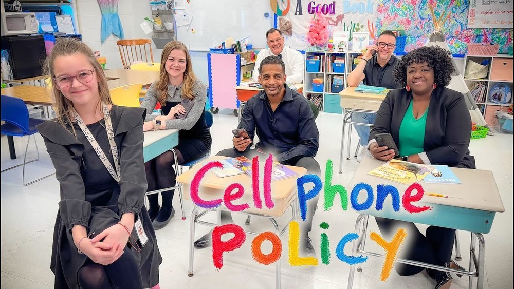 A group of six adults sit at colorful student desks in a classroom, each smiling and holding a cell phone. Behind them, the classroom is decorated with bright posters, bookshelves, and student artwork. The text "Cell Phone Policy" is written in crayon-style font across the image in various colors.