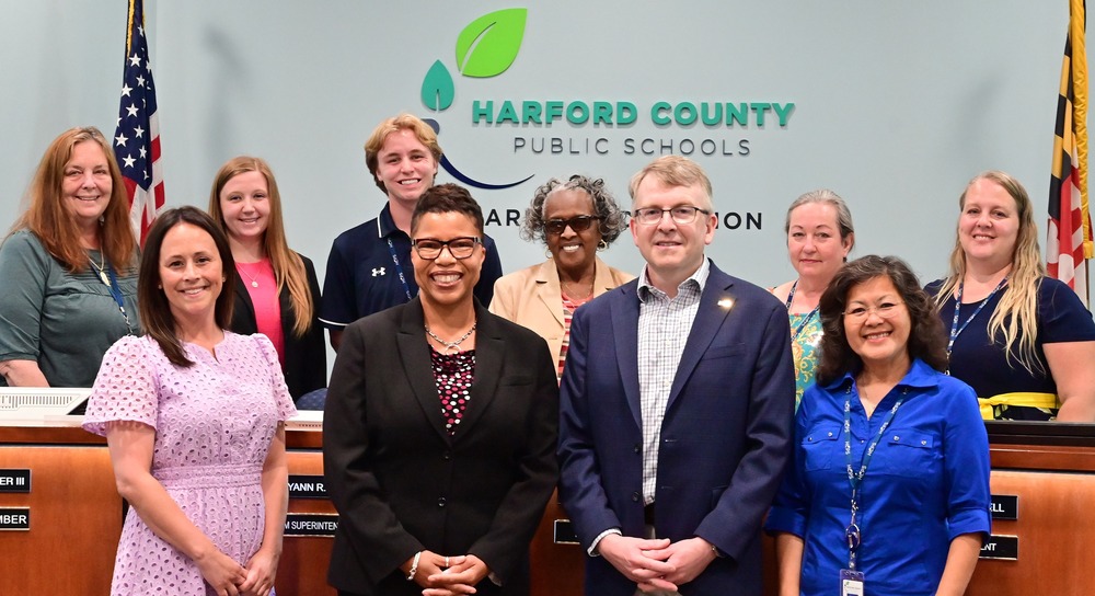 Board of education members stand with Dr. Mack in the board room in front of a sign that reads "Harford County Public Schools"