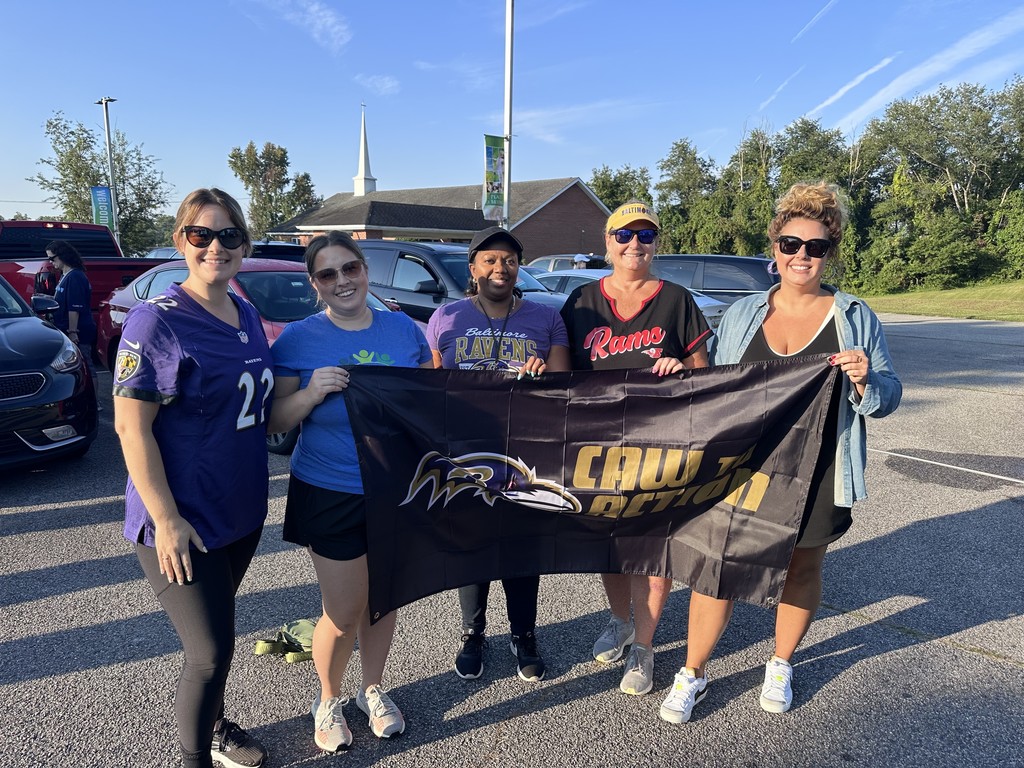 Five women standing in a parking lot holdin a ravens banner