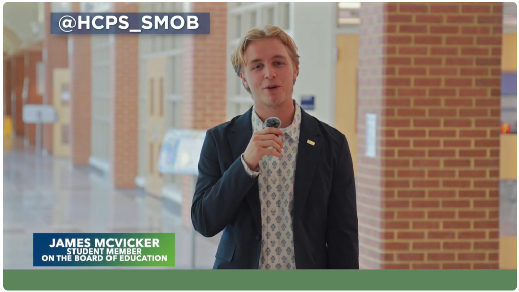 A student speaks into a microphone in a school hallway. On-screen text reads “@HCPS_SMOB” and “James McVicker, Student Member on the Board of Education.”