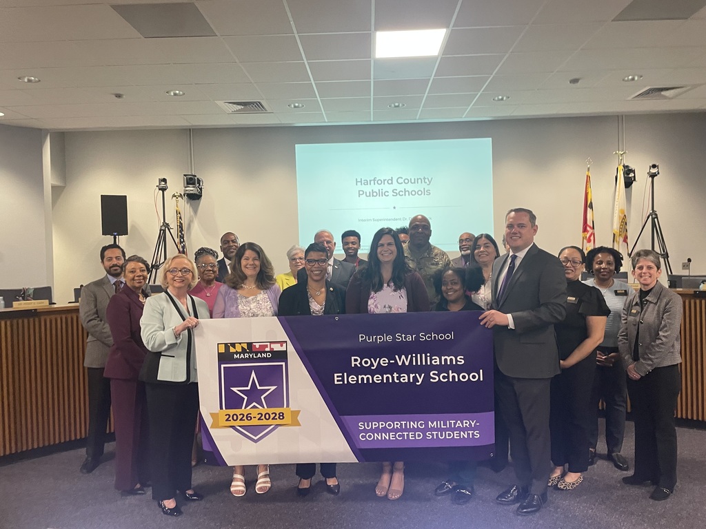 Group of individuals standing together in a board room holding a banner that shows Roye-Williams Elementary School as a Purple Star School