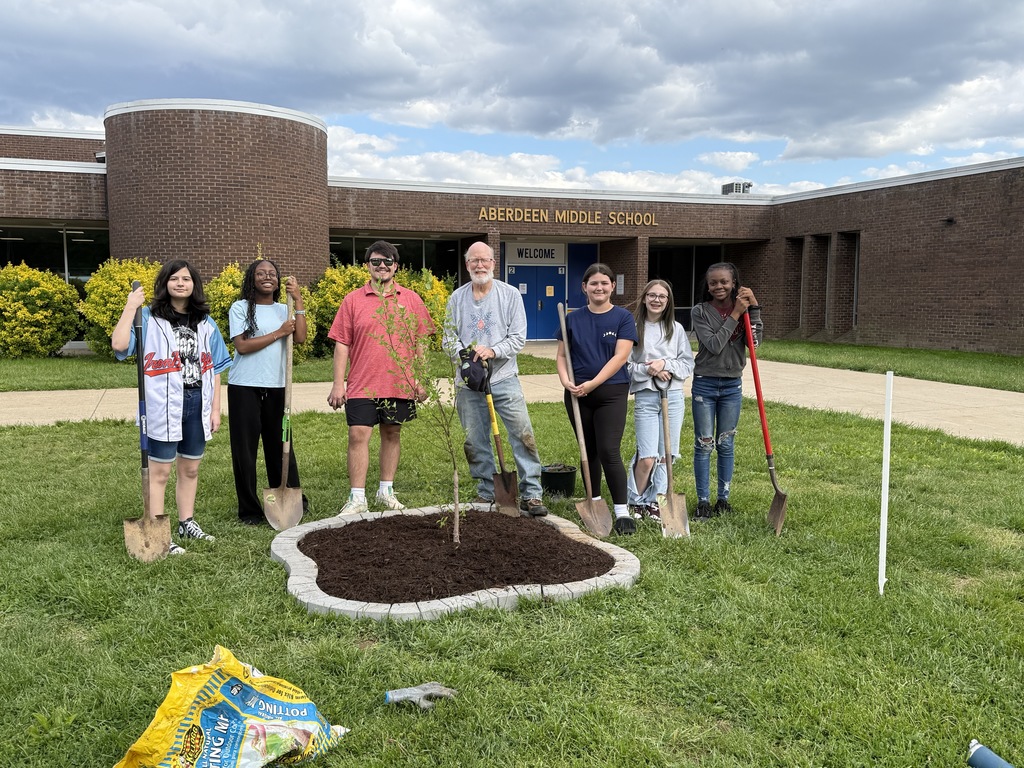 Green grass with teacher and students standing near a tree they planted.  Shows the green net and brown soil 