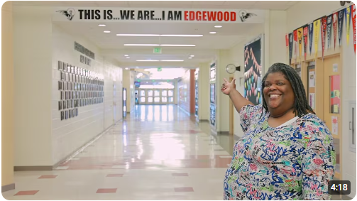 An image of Ms. Webster, and administrative assistant at Edgewood High School, in the hallway of EHS pointing as a sign that says, "This is...We are...I am Edgewood".