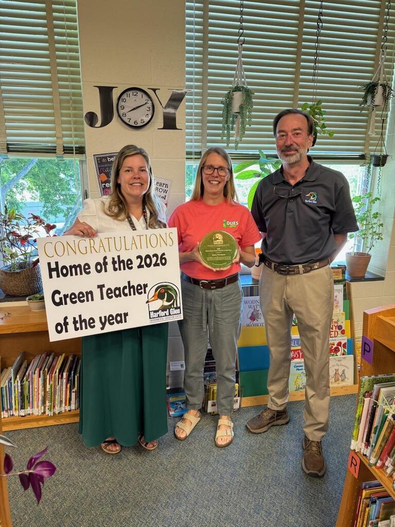 Three adults stand side by side in a classroom library area. The person on the left, labeled Mrs. Covelly, holds a large sign that reads, “CONGRATULATIONS Home of the 2026 Green Teacher of the year – Harford Glen.” The person in the middle, labeled Mrs. Thompson, holds a round green award plaque. The person on the right, labeled Mr. Eakes, stands with hands clasped. Behind them are bookshelves filled with children’s books, potted plants, hanging greenery, and windows with blinds.