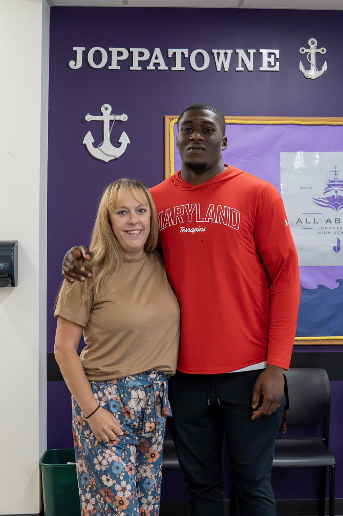 A woman in a tan top and floral pants smiles beside a young man in a red "Maryland Terrapins" hoodie, standing in front of a purple "Joppatowne" wall with nautical anchor decor and an "All Aboard" poster.