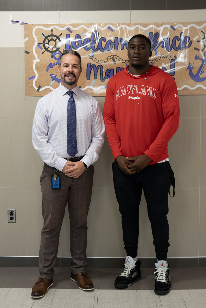 A man in a light checked dress shirt and tie stands beside a young man in a red "Maryland Terrapins" hoodie, in front of a nautical-themed "Welcome Back" banner featuring a ship's wheel and anchor.