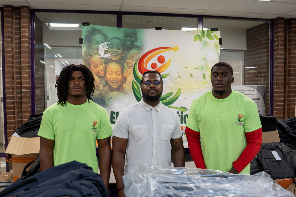 Three men stand in front of an "Elee Foundation" banner featuring smiling children. Two wear bright green Elee Foundation T-shirts, one with red long sleeves underneath, and the man in the middle wears a white button-up shirt. Backpacks and boxed supplies are visible on either side, suggesting a donation or backpack giveaway event.