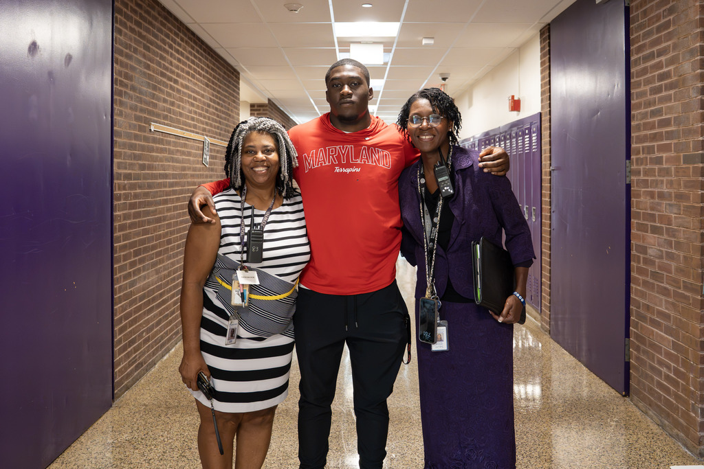 A young man in a red "Maryland Terrapins" shirt stands smiling between two school staff members in a hallway lined with purple lockers, his arms around their shoulders. The woman on the left wears a black-and-white striped dress with a staff radio, and the woman on the right wears a purple blazer