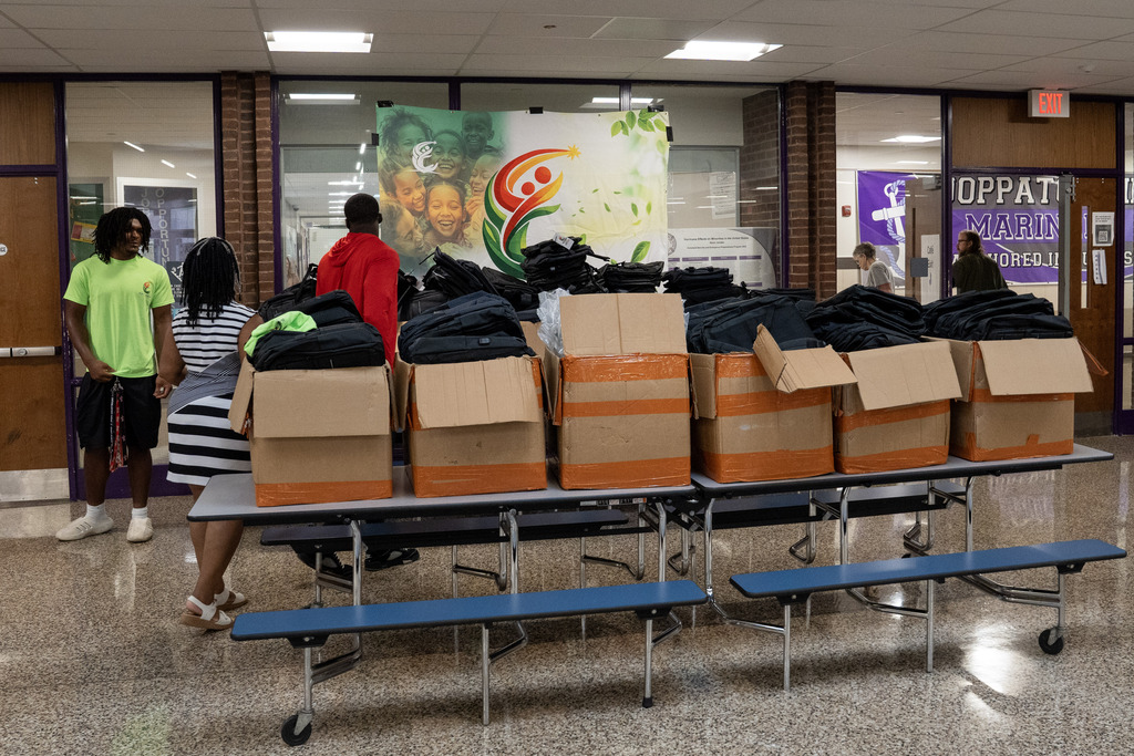 A long cafeteria table lined with open cardboard boxes full of backpacks sits in a school hallway, with a few people standing nearby and an "Elee Foundation" banner of smiling children hanging on the wall behind. A purple "Joppatowne Mariners" banner is visible on the far wall near an exit door.