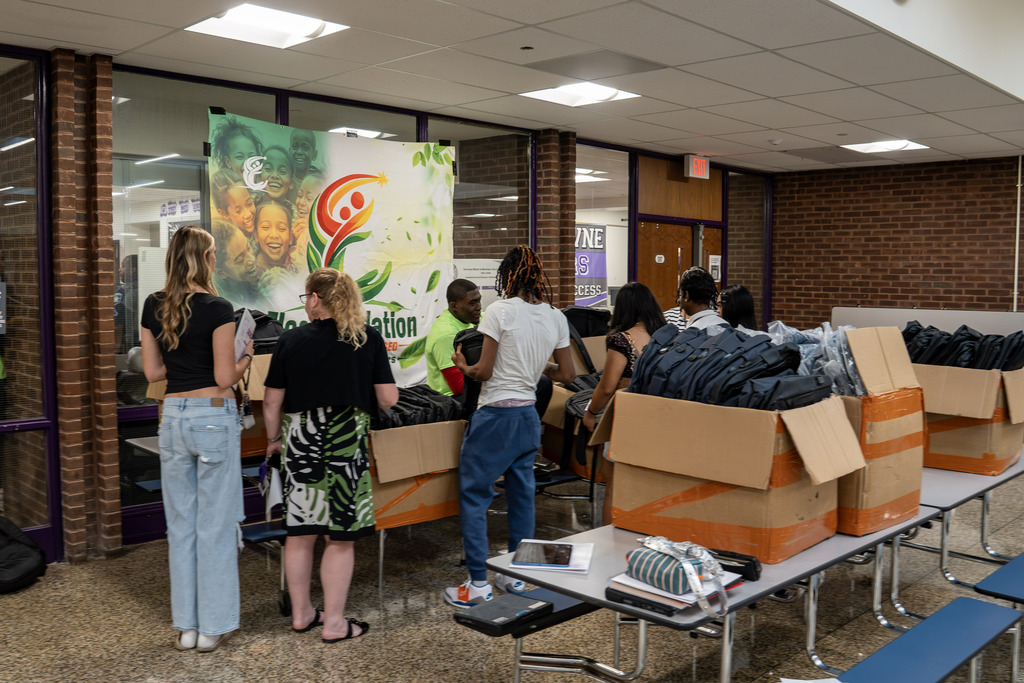 A group of people gather around tables lined with cardboard boxes full of backpacks, sorting and distributing them in a school hallway. An "Elee Foundation" banner with smiling children hangs on the wall behind them