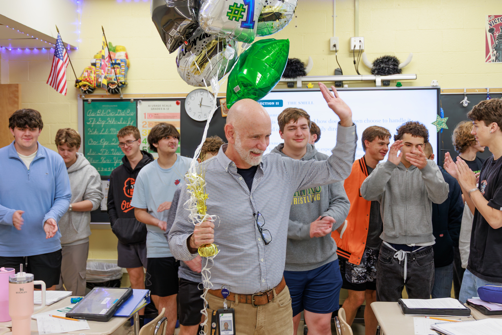 teacher standing in front of students being applauded and waiving while holding baloons.