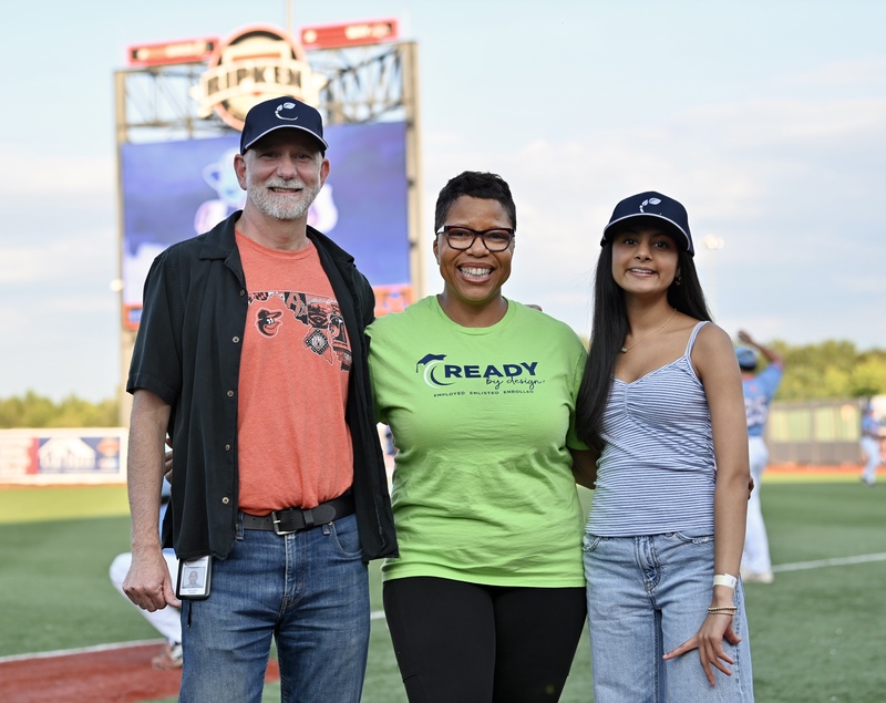 Three people stand smiling with a baseball scoreboard behind them