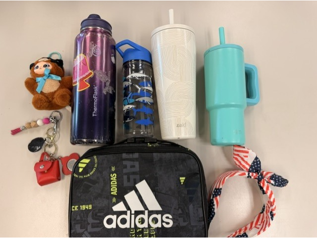 Overhead photo of several personal items arranged on a table, including an Adidas bag, four reusable water bottles and tumblers in purple, black, white, and teal, a red-white-and-blue hat, keys, a small red pouch, and a plush toy