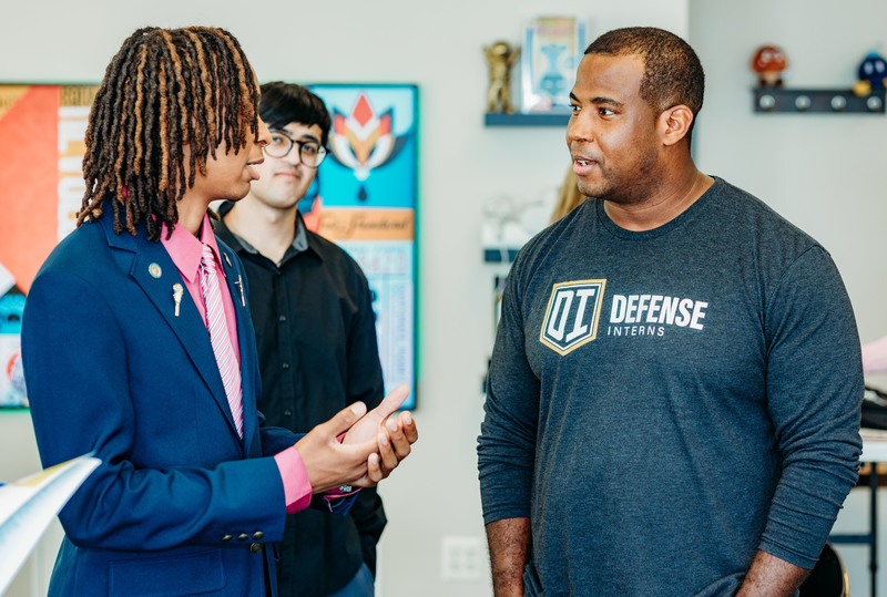 Adult wearing a Defense Interns shirt speaking with a student