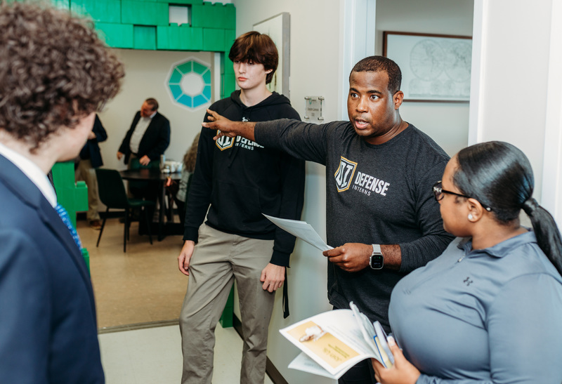 Adult wearing a Defense Interns shirt speaking with a group of students