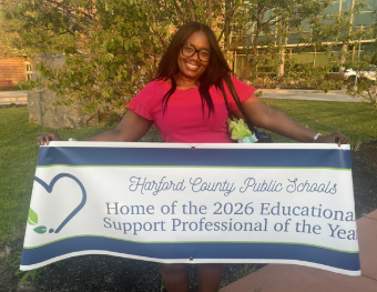 Ms. Laurence stands outside holding a banner that says Home of the 2026 Educational Support Professional of the Year.