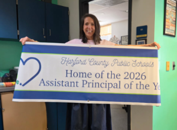 Ms. Sieracki stands in the main office at Bakerfield Elementary School holding a banner that says Home of the 2026 Assistant principal of the Year.