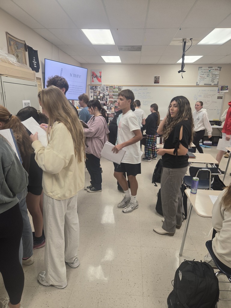People standing in a hallway, some near a screen, others near a table with chairs.