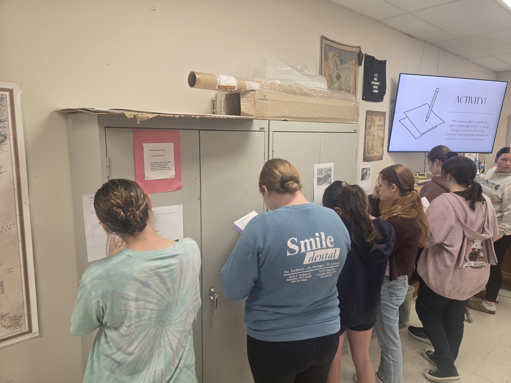 Several students stand in line facing a bulletin board. One student reads a paper in their hands.
