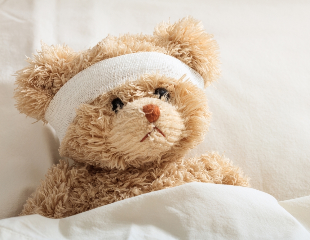 A teddy bear with a bandage around its head is tucked into bed.