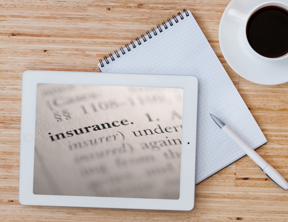 Tablet on a desk showing the word “insurance” on its screen, with a notebook, pen, and cup of coffee beside it.