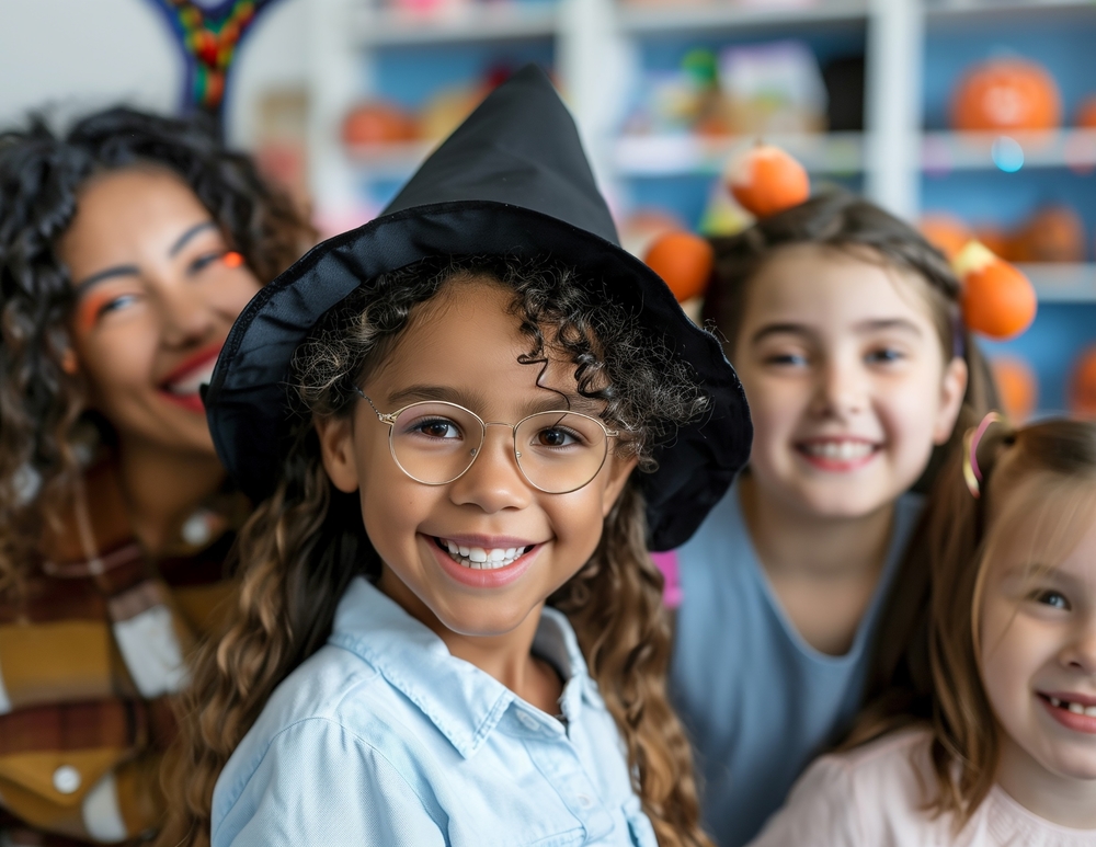 Four children smile at the camera, one wearing a witch hat and glasses, suggesting a Halloween celebration.