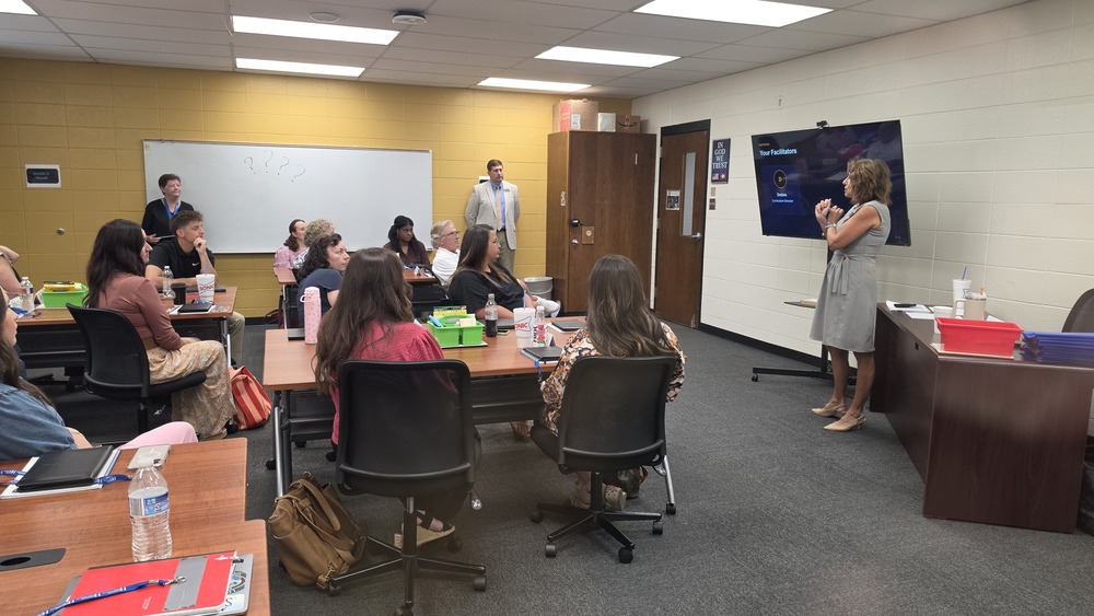 Mrs. Debbie Wilson, Director of Curriculum and Instruction, presents to a room of new teachers seated at tables during an inservice session at Harrison Schools Professional Development Center, while Superintendent Dr. Stewart Pratt observes in the background.