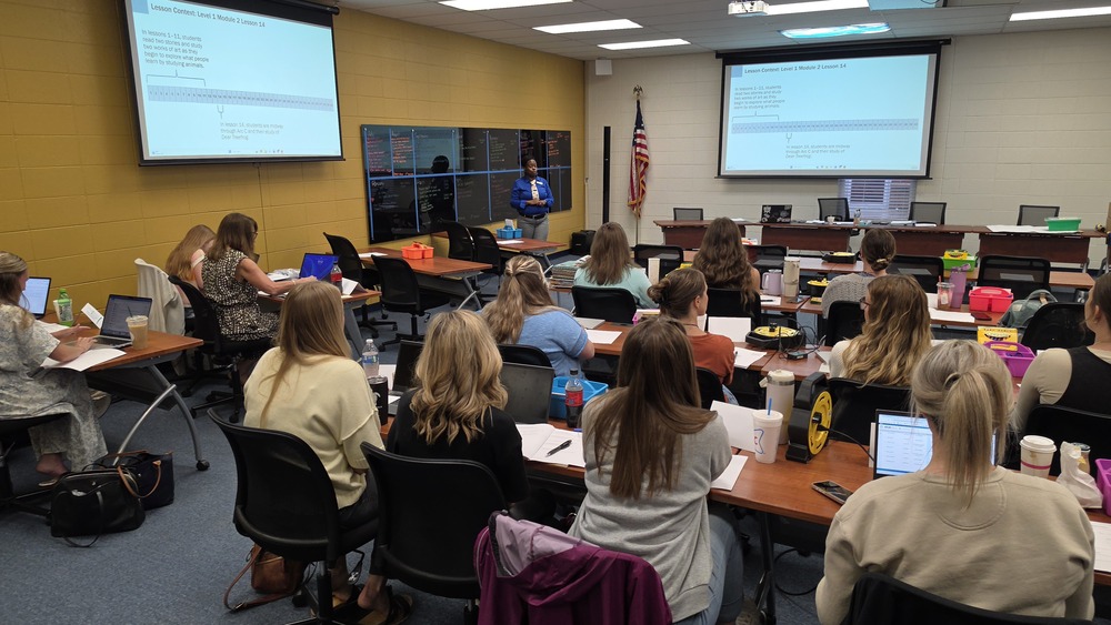 An indoor photo taken from the back of the Harrison School District professional development room. Approximately 15 teachers are seated at rows of long wooden tables, facing toward the front. Many have laptops, notebooks, and drinks open in front of them. At the front of the room, a female presenter in a blue shirt stands near a large black chalkboard. Two large projector screens on the wall display text and a timeline graphic titled "Lesson Context: Level 1, Module 2, Lesson 14." An American flag stands in the background corner.