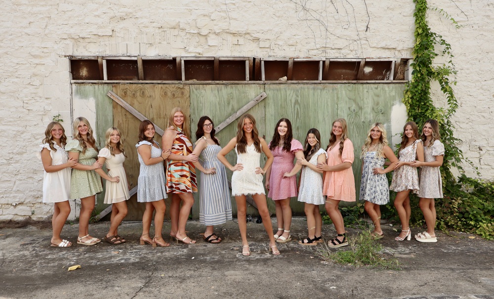 Group photo of thirteen young women representing Harrison High School's Homecoming court in short summer dresses, standing in a line outdoors against a rustic green wooden door and white brick wall. They are linked arm-in-arm, with Queen Ella Ramsey in a white lace dress standing confidently in the center.”