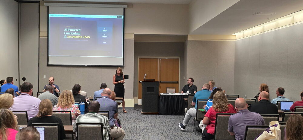 Harrison School District educators leading a workshop in a conference room at the ADE Summit. A female presenter in a black dress stands speaking near a large projection screen displaying a slide titled "AI-Powered Curriculum & Instruction Tools." Two co-presenters sit at a panel table nearby, while an audience of educators sits in rows of chairs watching the presentation.