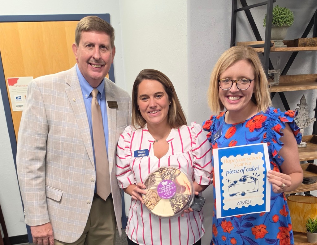 Three people standing together indoors smiling for the camera. On the left is a man in a beige plaid blazer and tan trousers. In the center, a woman wearing a white top with pink vertical stripes holds a round, multi-flavor cheesecake in a clear plastic container. On the right, a woman in a blue floral dress holds a sign that reads, "Hope your school year is a piece of cake!" with an Arvest logo at the bottom.