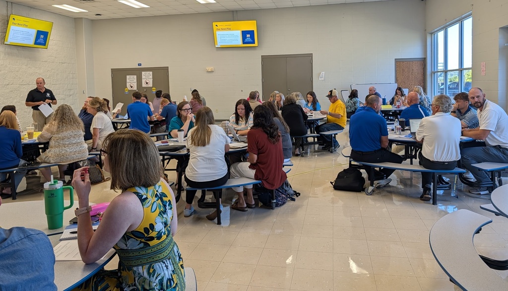 A large room filled with educators and staff seated at round cafeteria tables, working in groups with paperwork and laptops. A presenter stands near the wall on the left holding papers, while two wall-mounted TV monitors display presentation slides titled "Our Next Play." Bright sunlight filters in through large windows on the right.
