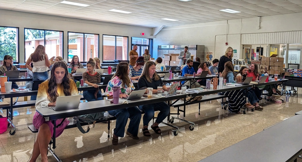 A wide room filled with teachers and school staff seated along long rows of folding tables, actively working on laptops and taking notes. Windows line the left wall, showing outdoor school buildings, while cardboard boxes and school equipment are stacked in the background on the right.