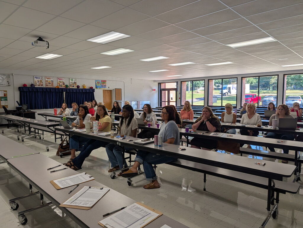 A group of teachers and school staff sitting at long cafeteria tables during an indoor training session or meeting, with laptops, clipboards, and notebooks open in front of them.