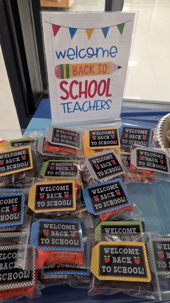 A framed sign sitting on a blue tablecloth reads "welcome BACK TO SCHOOL TEACHERS" in colorful lettering with a pencil illustration and colorful banner flags at the top. In front of the sign are several small, clear plastic treat bags containing candy bars. Each bag is sealed with a colorful gift tag styled like a chalkboard that says "WELCOME BACK TO SCHOOL!" flanked by red apples.