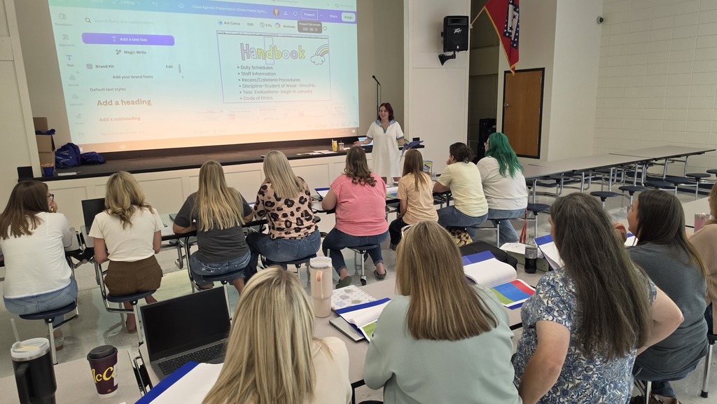 An audience of educators sitting at long tables, viewed from behind, watching a presenter stand before a large projector screen. The screen shows a Canva presentation slide labeled "Handbook" covering topics like duty schedules, staff information, and cafeteria procedures. An Arkansas state flag hangs near a stage doorway on the right.