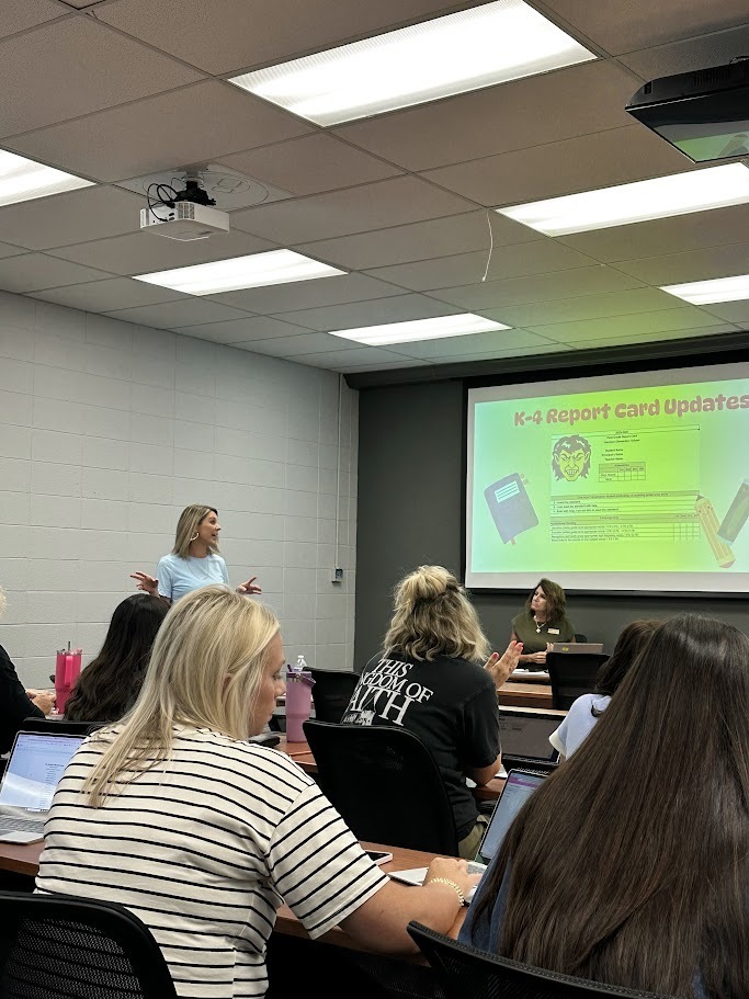 A presenter stands on the left side of a classroom speaking to a seated audience of teachers with open laptops. A large projector screen displays a slide titled "K–4 Report Card Updates" showing a sample report card graphic with the Harrison Schools mascot.