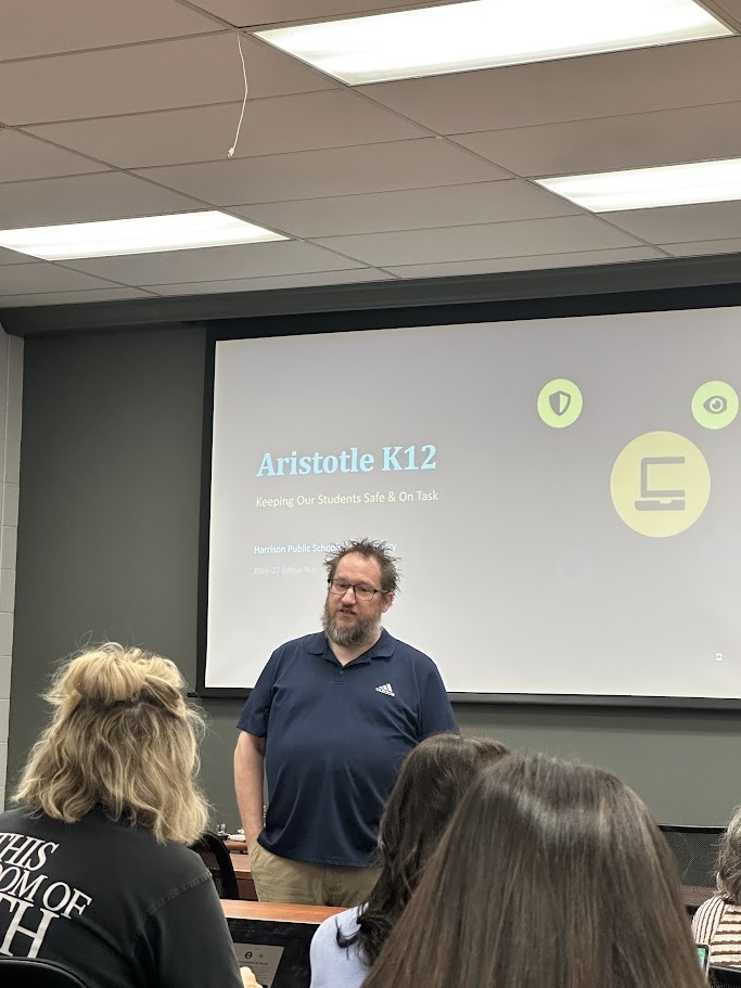 A presenter in a dark blue polo shirt stands at the front of a room speaking to teachers. Behind him, a projection screen reads "Aristotle K12: Keeping Our Students Safe & On Task" with icons representing web safety and monitoring.