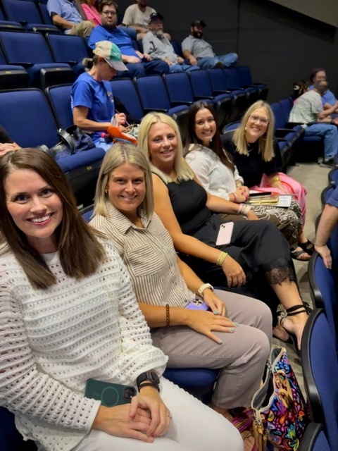 A close-up shot of five smiling female teachers sitting together in a row of auditorium seats, looking up at the camera before the presentations begin.